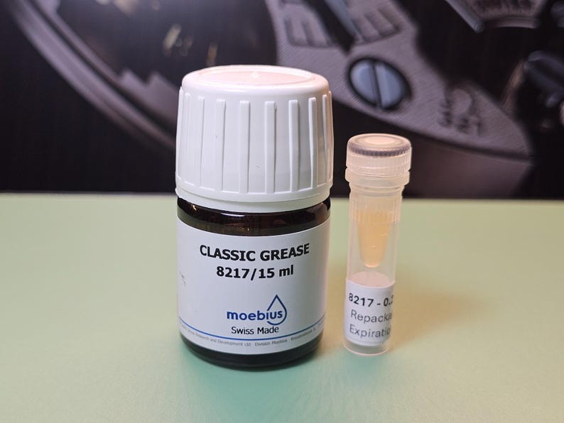 May include: A small glass bottle of "CLASSIC GREASE 8217/15 ml" with a white cap and a smaller clear vial containing a yellow liquid. The label reads "moebius Swiss Made". The background is a blurred image of a watch.