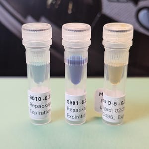 May include: Three clear vials with white caps, each containing a different colored liquid: light blue, dark blue, and yellow. Each vial has a white label with text including "Repackaged" and "Expiration".