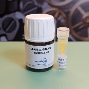 May include: A small, dark glass bottle with a white cap labeled "CLASSIC GREASE 8200/15 ml" and a smaller clear vial containing a yellow liquid. The bottle has a blue logo and the words "moebius Swiss Made".