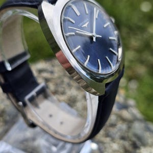 May include: A close-up of a vintage wristwatch with a dark blue face and silver hands. The watch has a black leather band and a silver case. The word "LONGINES" is printed on the watch face.