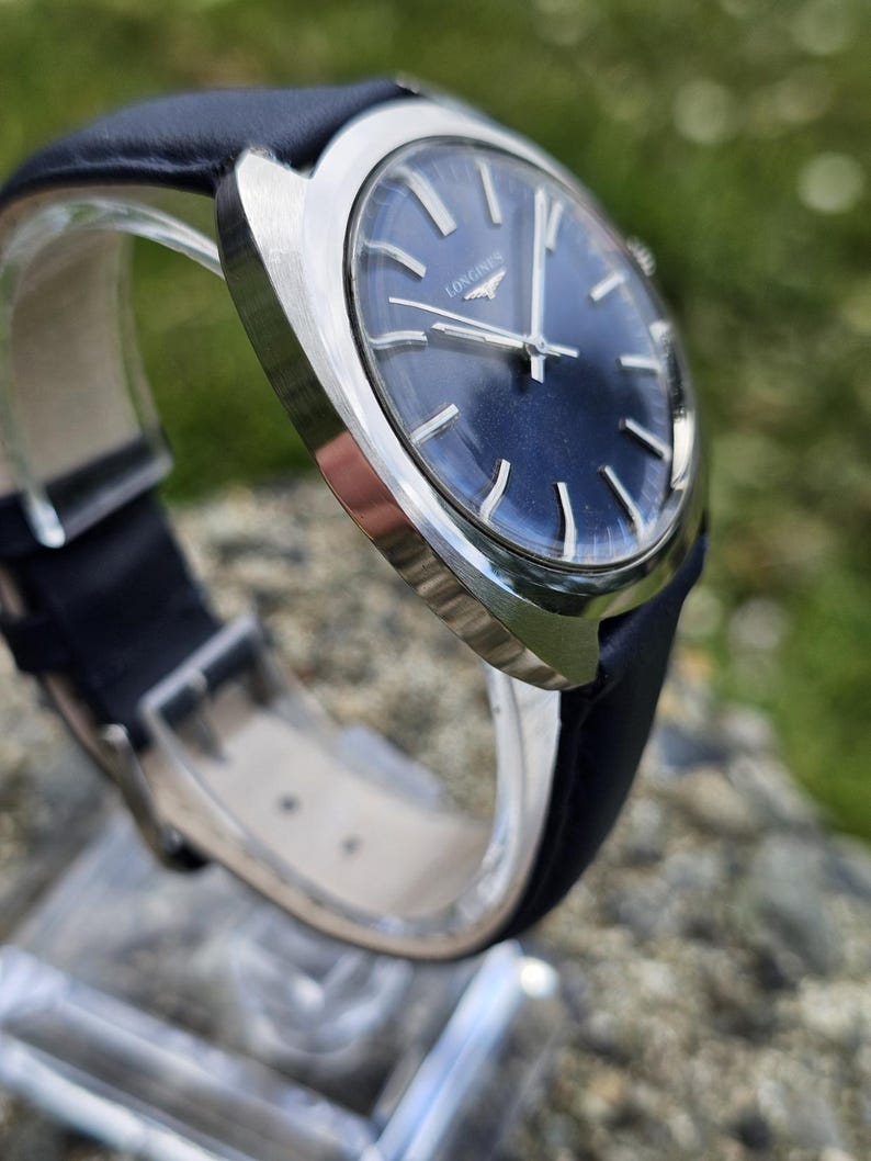 May include: A close-up of a vintage wristwatch with a dark blue face and silver hands. The watch has a black leather band and a silver case. The word "LONGINES" is printed on the watch face.