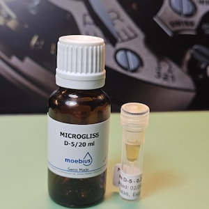 May include: A brown glass bottle with a white cap and a small clear vial. The bottle has a white label with the text "MICROGLISS D-5/20 ml" and the brand "moebius". The vial contains a yellow liquid.