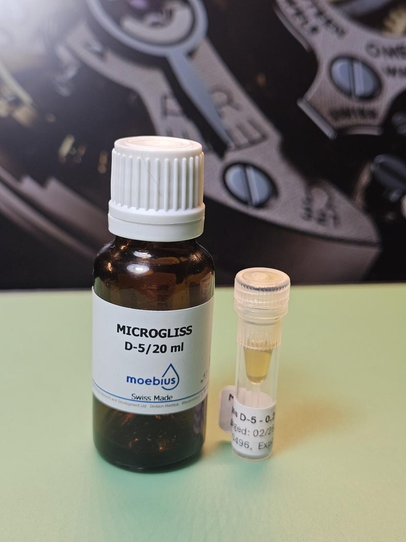 May include: A brown glass bottle with a white cap and a small clear vial. The bottle has a white label with the text "MICROGLISS D-5/20 ml" and the brand "moebius". The vial contains a yellow liquid.