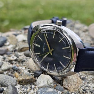 May include: A close-up of a vintage Longines watch with a dark blue face and silver casing. The watch has gold hands and markers, and a dark blue leather band. The watch is resting on a bed of small rocks.