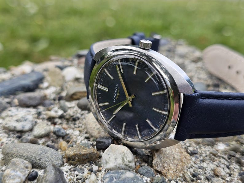 May include: A close-up of a vintage Longines watch with a dark blue face and silver casing. The watch has gold hands and markers, and a dark blue leather band. The watch is resting on a bed of small rocks.
