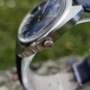 May include: A close-up of a silver wristwatch with a dark blue face and a blue leather band. The watch has a textured crown and a sleek, modern design. The watch face has silver hour markers.
