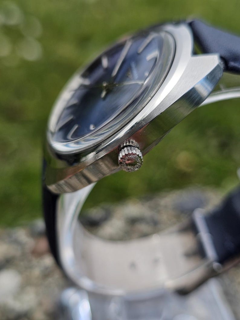 May include: A close-up of a silver wristwatch with a dark blue face and a blue leather band. The watch has a textured crown and a sleek, modern design. The watch face has silver hour markers.