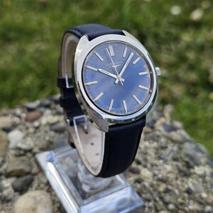 May include: A vintage wristwatch with a blue dial and silver-tone case. The watch features silver hands and markers, and a black leather strap. The watch is displayed on a clear stand, showcasing its classic design. The watch face has the word "Longines".