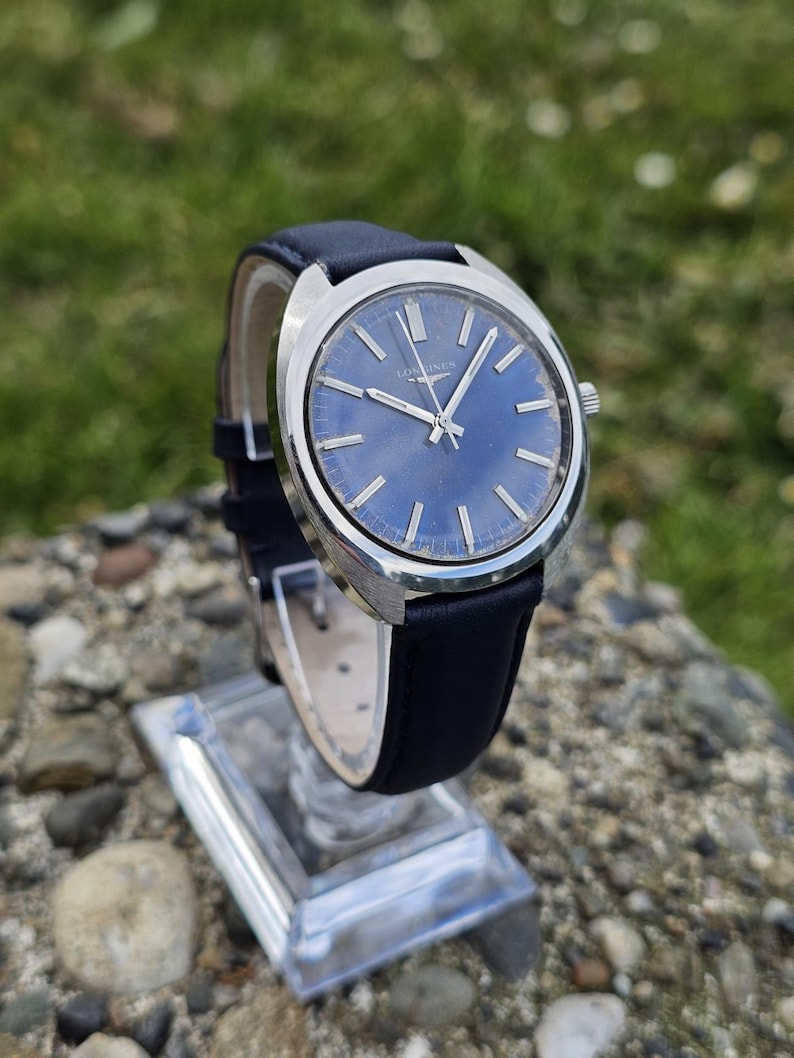 May include: A vintage wristwatch with a blue dial and silver-tone case. The watch features silver hands and markers, and a black leather strap. The watch is displayed on a clear stand, showcasing its classic design. The watch face has the word "Longines".