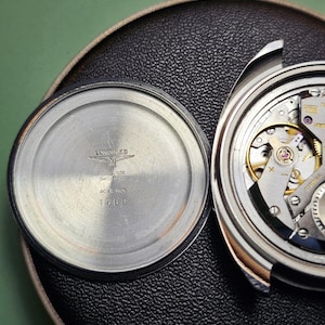 May include: Close-up of a disassembled Longines watch. The watch's back is silver with the brand name, logo, and "ACIER INOX 1500" text. The watch's inner workings are visible, showcasing intricate gears and mechanisms. The watch is on a black textured surface.