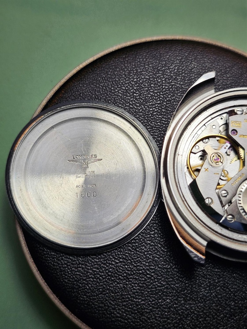 May include: Close-up of a disassembled Longines watch. The watch's back is silver with the brand name, logo, and "ACIER INOX 1500" text. The watch's inner workings are visible, showcasing intricate gears and mechanisms. The watch is on a black textured surface.