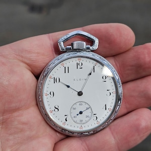 May include: A silver-toned Elgin pocket watch with a white face and black numerals. The watch has a small seconds dial and ornate detailing around the bezel. The watch is held in a hand.