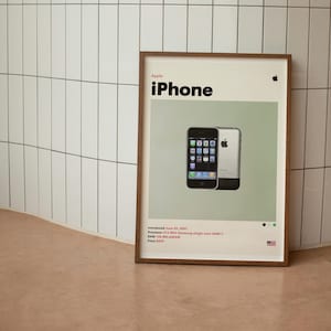 Puede incluir: Un póster enmarcado que presenta el primer iPhone. El póster muestra el logotipo de Apple y la palabra "iPhone" en negrita negra. Debajo del título hay una imagen del teléfono. La impresión incluye especificaciones técnicas y la fecha de introducción.