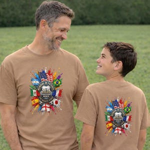 Peut inclure: Deux t-shirts beiges avec un graphisme coloré. Le motif comprend des drapeaux de différents pays, des ballons de football et le texte "Mundial de Futebol 2026 Soccer World The Biggest Stage".