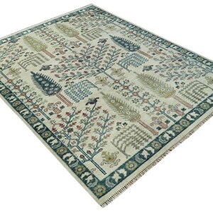 Puede incluir: Una alfombra beige con un patrón repetitivo de árboles estilizados en tonos de verde, marrón y rojo. La alfombra tiene un borde azul y verde con flecos.