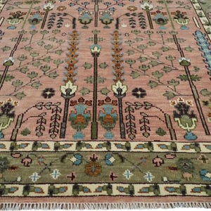 May include: A detailed, close-up view of a rug with a pinkish-red background. The rug features a repeating pattern of stylized trees and floral motifs in shades of green, blue, and brown. The border is a darker green with geometric designs.