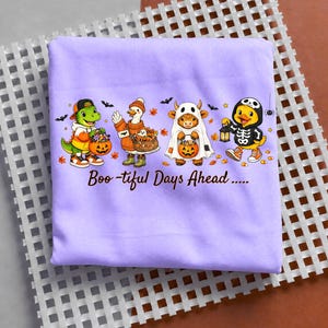 May include: A light purple t-shirt featuring a graphic of a dinosaur, a goose, a highland cow, and a duck dressed in Halloween costumes, each holding a jack-o'-lantern bucket. The text below reads Boo-tiful Days Ahead with small bats.