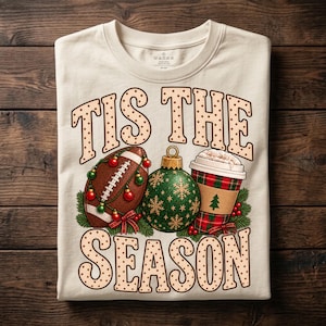 Peut inclure: Un t-shirt couleur crème avec le texte TIS THE SEASON en lettres marron à pois. Le motif comprend un ballon de football américain entouré de boules rouges et vertes, une boule de Noël verte à flocons dorés et un gobelet à café à motif écossais.