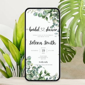 May include: A smartphone displays a bridal shower invitation. The invitation features the words "bridal shower" in elegant script, along with the bride's name, date, and time. Eucalyptus and other greenery frame the text. The phone is set against a backdrop of green plants.