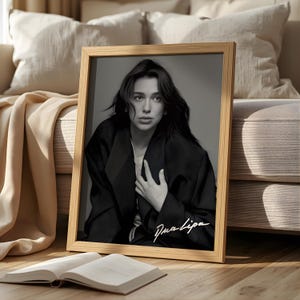 Può includere: Un ritratto in bianco e nero incorniciato di Dua Lipa. L'immagine la mostra con un blazer nero, con il suo nome in uno stile di firma in basso a destra. Un libro aperto è appoggiato davanti alla cornice, con uno sfondo dai toni neutri.