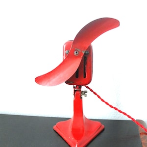 May include: A vintage red electric fan with a single, curved blade. The fan has a rectangular motor housing and a triangular base. A red, twisted power cord extends from the back. The fan is likely from the mid-20th century.