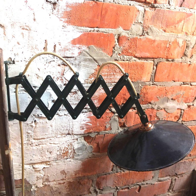 May include: A black metal wall-mounted lamp with an accordion-style arm and a black metal shade. The lamp is mounted on a brick wall.
