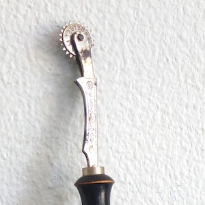 May include: A silver sewing tool with a black wooden handle. The tool has a wheel with numbers 1-28 around the edge and a decorative design on the handle.
