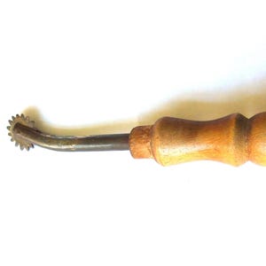 Pode incluir: Uma ferramenta de costura vintage, uma roda de traçar, com um cabo de madeira e uma roda de metal com pequenos dentes. O cabo é castanho claro, e os componentes metálicos mostram sinais de idade.