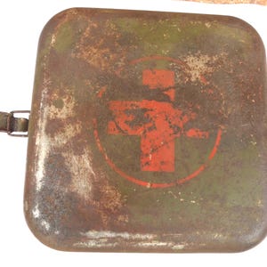 Op de afbeelding: Een vierkante, metalen vintage EHBO-kit met een roestig, verweerd uiterlijk. De kit is olijfgroen met een rood kruis embleem in het midden. Een metalen sluiting en scharnier zijn zichtbaar aan de zijkant. De algehele esthetiek suggereert een gebruikt, antiek item.