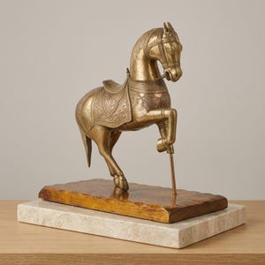 Può includere: Una scultura di cavallo in ottone in posa rampante, caratterizzata da intricati motivi floreali incisi sulla sella e sul corpo. Il cavallo è sostenuto da un'asta metallica sottile fissata su una base in legno rettangolare, poggiata su una lastra di marmo.