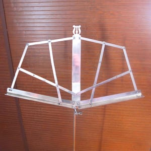 May include: A silver-colored, adjustable music stand. The stand features a lyre-shaped design at the top and a wide, angled shelf for holding sheet music. The stand is made of metal and has a telescoping pole for height adjustment.