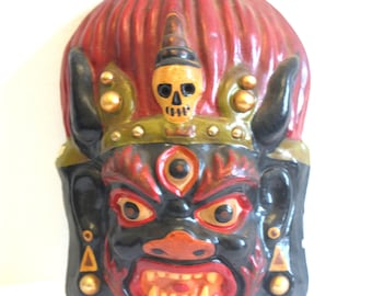 May include: A hand-painted wooden mask with a black face, red and yellow details, and a skull on the forehead. The mask has a third eye and horns.