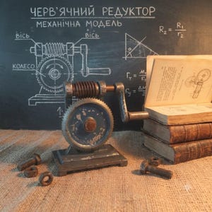 May include: A mechanical worm gear model featuring a grey metal base, a central gear, and a hand crank. The device is positioned near a stack of old books and loose metal bolts. The background is a chalkboard with technical diagrams and text.