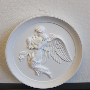 May include: White plaster relief sculpture of an angel with wings holding a small child. The angel is depicted with flowing drapery and a gentle expression.