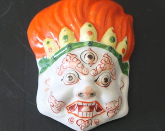 May include: A white ceramic mask with a red, yellow, and green painted design. The mask has a third eye and a fierce expression.