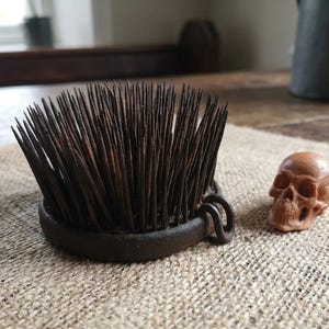 May include: A circular metal flower frog featuring numerous upright, dark, pointed needles set into a heavy base with two small rings on the side, accompanied by a small, light brown, carved wooden skull figurine.