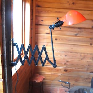 May include: An orange metal desk lamp with a black articulated arm, mounted on a wooden wall. The lamp has a classic design with an adjustable arm and a bright orange shade. The lamp is attached to the wall with a black accordion-style arm.