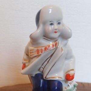 以下が含まれることがあります： 青と白の帽子とコートを着た子供の磁器製フィギュア。子供は手に赤と白の物を持っています。フィギュアは金色のアクセントで描かれています。