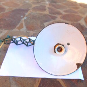 May include: A white metal lampshade with a black rim and a yellow light bulb socket. The lampshade is attached to a black metal frame with a series of square shapes.