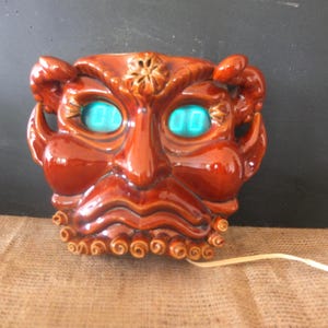Puede incluir: Un reloj de cerámica esmaltada marrón con forma de rostro con cuernos y expresión ceñuda. El reloj presenta dos pantallas digitales de color turquesa brillante como ojos, un detalle floral central en la frente y una barba rizada en el borde.