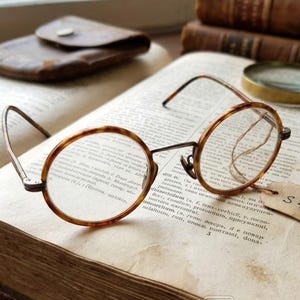 Peut inclure: Un livre ancien ouvert avec des lunettes rondes, une loupe et un étui en cuir. Les lunettes ont un motif écaille de tortue. Une petite étiquette avec le numéro "S 401" est attachée au livre.