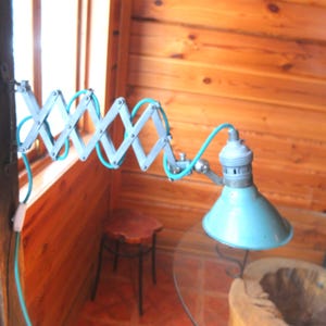 Könnte beinhalten: Verstellbare Vintage-Wandleuchte mit hellblauem Schirm und Ziehharmonika-Arm. Die Lampe ist an einer Holzwand befestigt. Ein kleiner Holzstuhl und ein Glastisch sind im Hintergrund zu sehen. Der Arm misst ca. 60 cm.