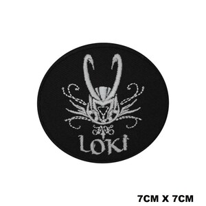 May include: A circular black embroidered patch measuring 7cm by 7cm. The design features a white stitched horned helmet emblem centred above the text LOKI in a stylised font, with decorative swirling lines extending from the sides of the helmet.