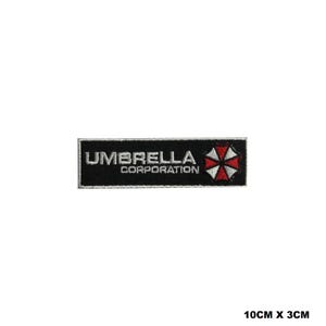 May include: A rectangular embroidered patch with a black background and white border. The text reads UMBRELLA CORPORATION in white capital letters. To the right is a circular logo divided into eight red and white segments. Dimensions are 10cm x 3cm.