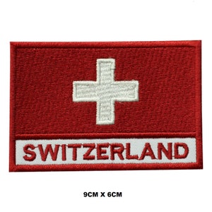 Puede incluir: Un parche bordado rojo y blanco con la bandera suiza y la palabra "SUIZA" en letras blancas. El parche mide 9 cm por 6 cm.