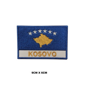 May include: Embroidered patch featuring the flag of Kosovo. The patch is blue with a gold outline and six white stars. The word "KOSOVO" is written in gold below the flag.