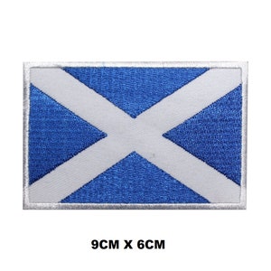 May include: An embroidered patch featuring the flag of Scotland, in blue and white. The patch is 9 cm by 6 cm and has a white border.