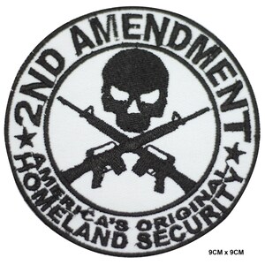 May include: A white circular patch with black embroidery. The patch features a skull and crossbones with two rifles. The text "2nd Amendment" is written around the top and bottom of the circle. The text "America's Original Homeland Security" is written around the bottom of the circle.