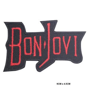 Puede incluir: Un parche bordado negro con el texto rojo BON JOVI en una fuente estilizada. El parche tiene una forma distintiva con una extensión vertical en la letra J. Las dimensiones de 9cm x 4.5cm están claramente impresas en la imagen.