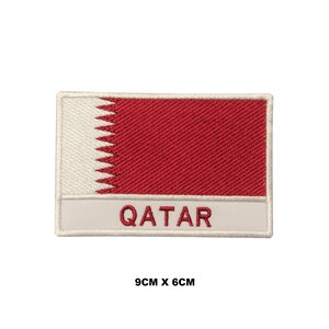 Puede incluir: Parche bordado blanco y rojo con la palabra "QATAR" en letras blancas. El parche mide 9 cm por 6 cm.